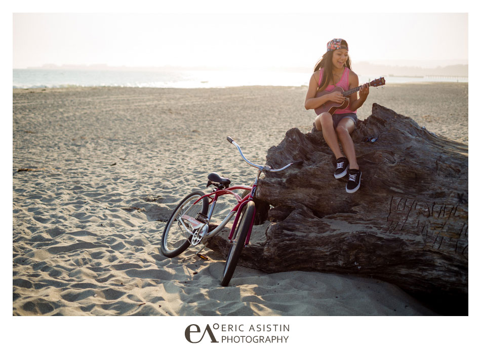 high school senior plays her ukelele on the beach sitting on large driftwood with her beach cruiser